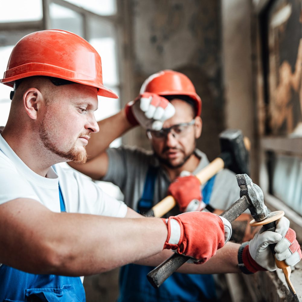Two workers at a very big construction site removing plaster from an old brick wall near a glass window
