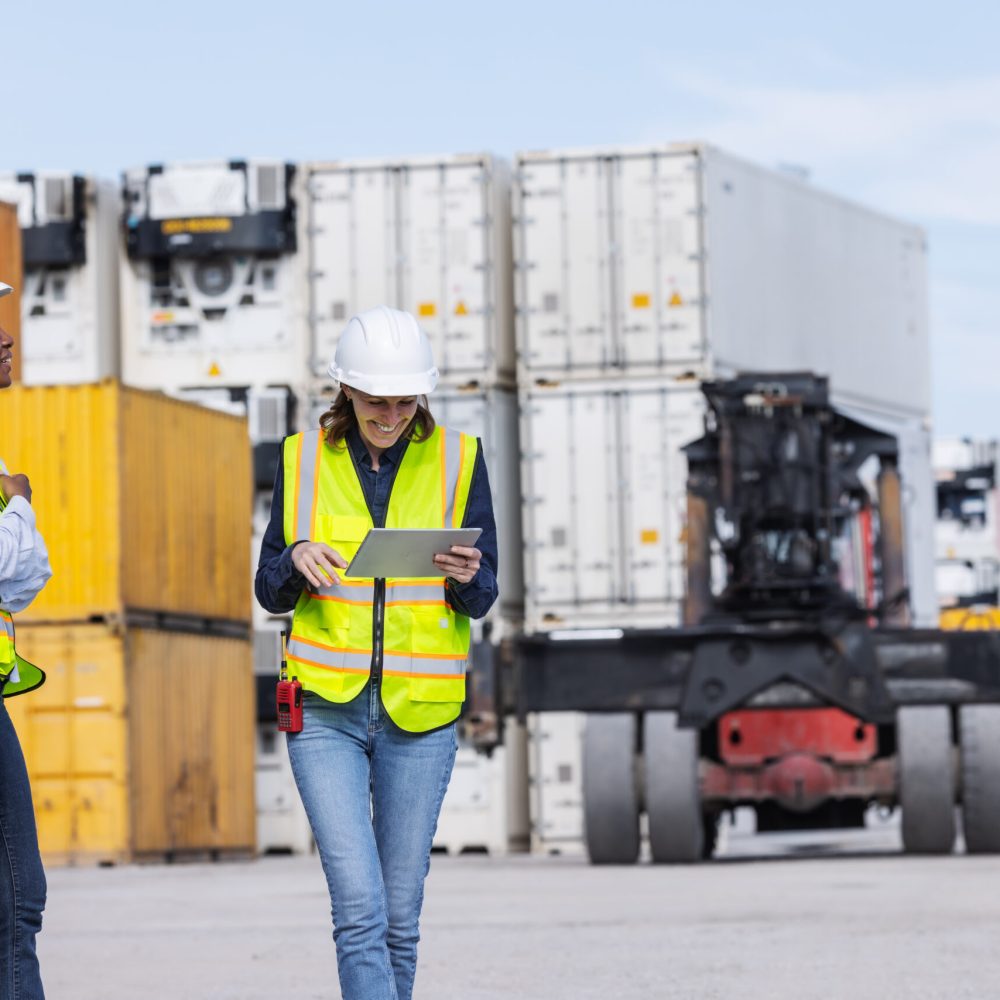 Two female workers in safety vests and hard hats discuss operations at a shipping yard, one holding a tablet. Stacked containers and heavy machinery highlight logistics, teamwork, and global trade.